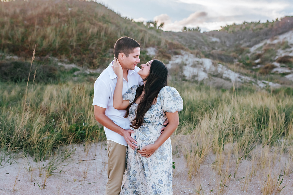 Couple in love embraces in grassy sand dunes. Woman holds her belly in a floral dress; man in white shirt smiles at her. Calm, romantic setting.