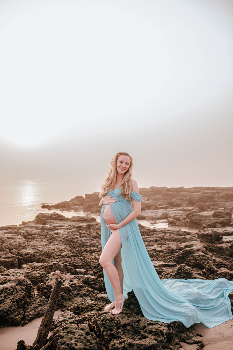 Pregnant woman in a flowing blue dress smiles, standing on rocky shore at sunset. Calm sea and bright sky create a serene backdrop.
