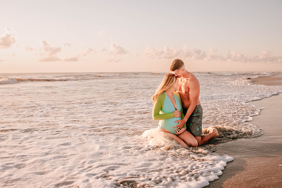 Couple kneeling in shallow ocean waves at sunset. Woman in green swimwear holds her belly; man embraces her warmly. Serene beach scene.