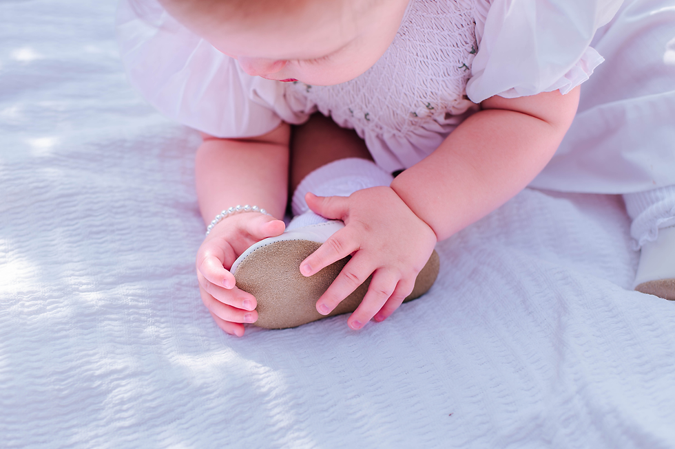 Baby wearing white clothes, sitting on a textured white blanket, holding a shoe during her milestone family photos. Soft lighting and pastel colors create a gentle mood.