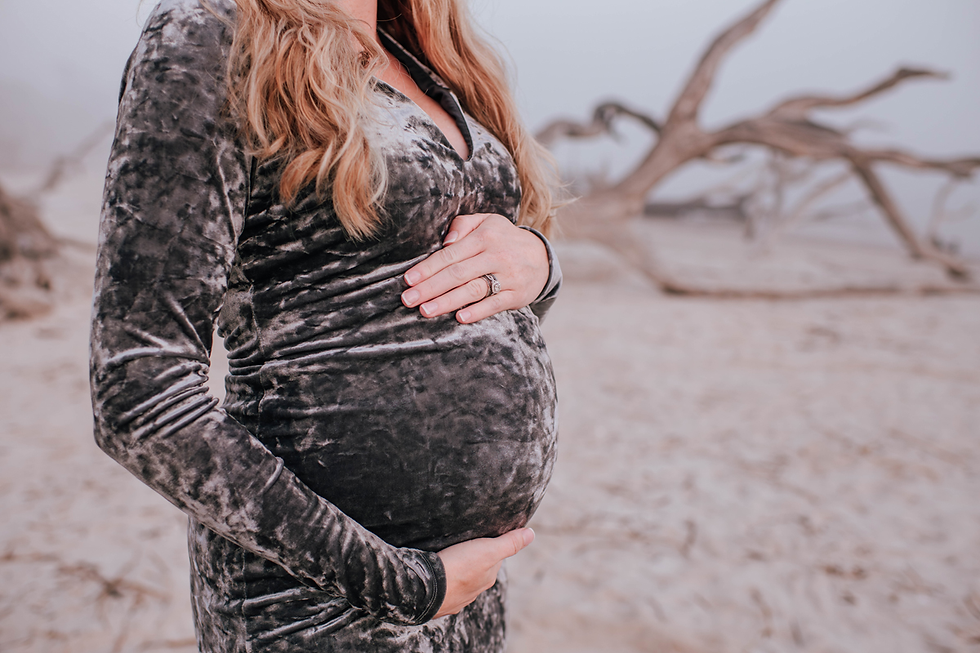 Pregnant woman in gray dress cradles her belly on a sandy beach with driftwood. Calm, serene setting with muted colors.