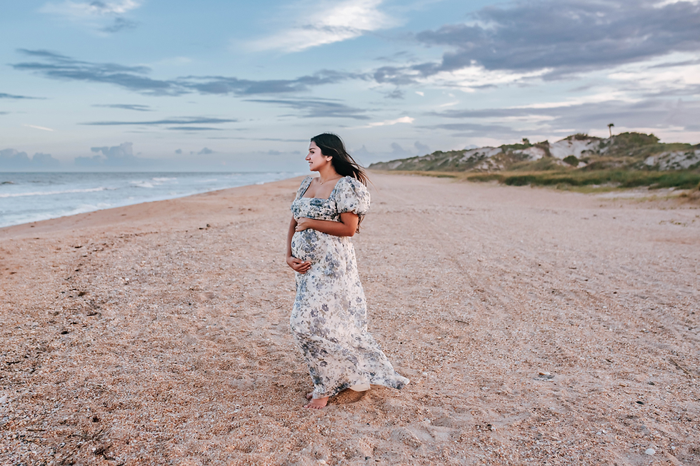 Pregnant woman in a floral dress stands on a sandy beach, gazing at the ocean. The sky is partly cloudy, creating a serene atmosphere.