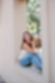 Smiling woman in blue jeans and a white top sits on a window ledge during her high school senior portrait session. Sunlit background with green trees. Relaxed, cheerful mood.
