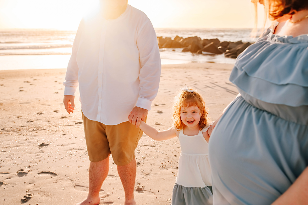 Pregnant woman, man, and smiling child hold hands on a sunlit beach. The woman wears a blue dress; ocean waves are in the background.