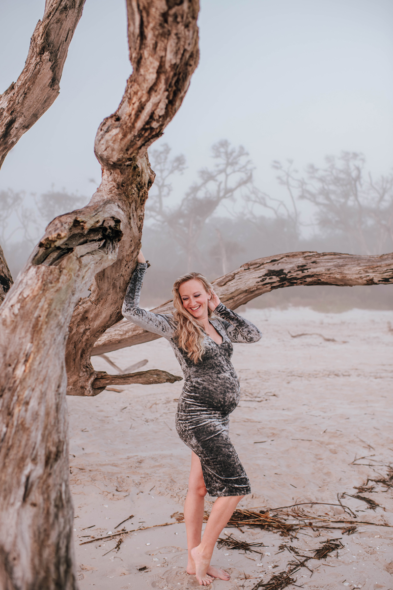 Pregnant woman in a gray dress poses happily against a weathered tree on a misty beach, surrounded by driftwood.