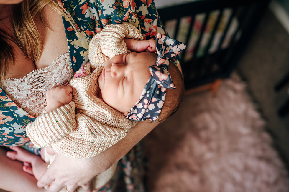 Mother in a floral dress holds a sleeping baby in a knitted outfit with a floral headband. Background includes a crib and pink rug.
