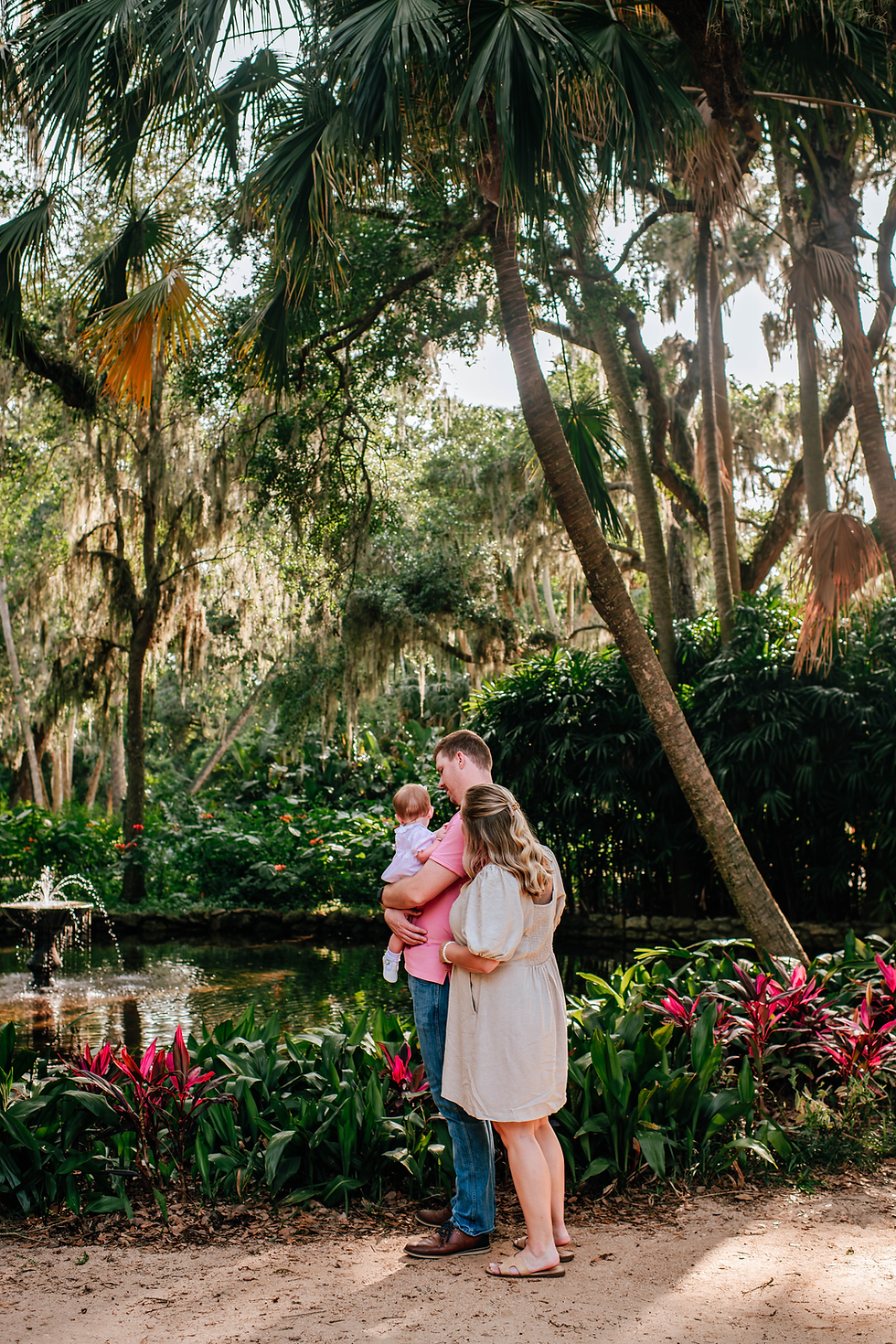 Family of three stands near a pond in a lush garden. Sunlight filters through palm trees, highlighting vibrant red and green foliage.