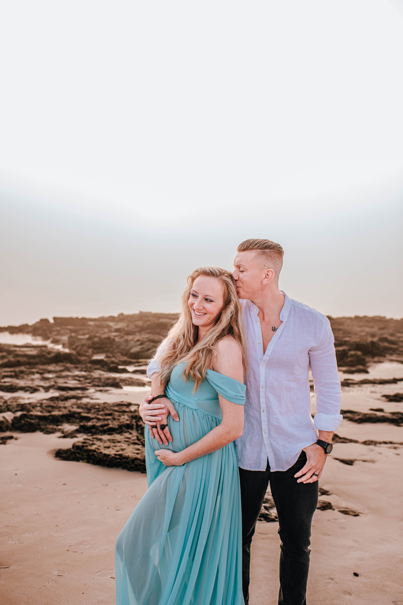 A couple on a beach; the man kisses the woman's forehead. She's in a flowing teal dress, smiling. Rocky sand background, serene mood.