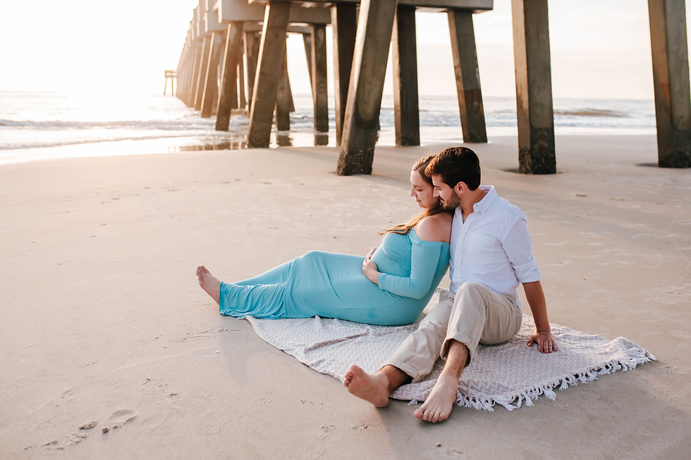 expecting couple sitting on blanket cuddling together on beach with ocean sunrise behind them