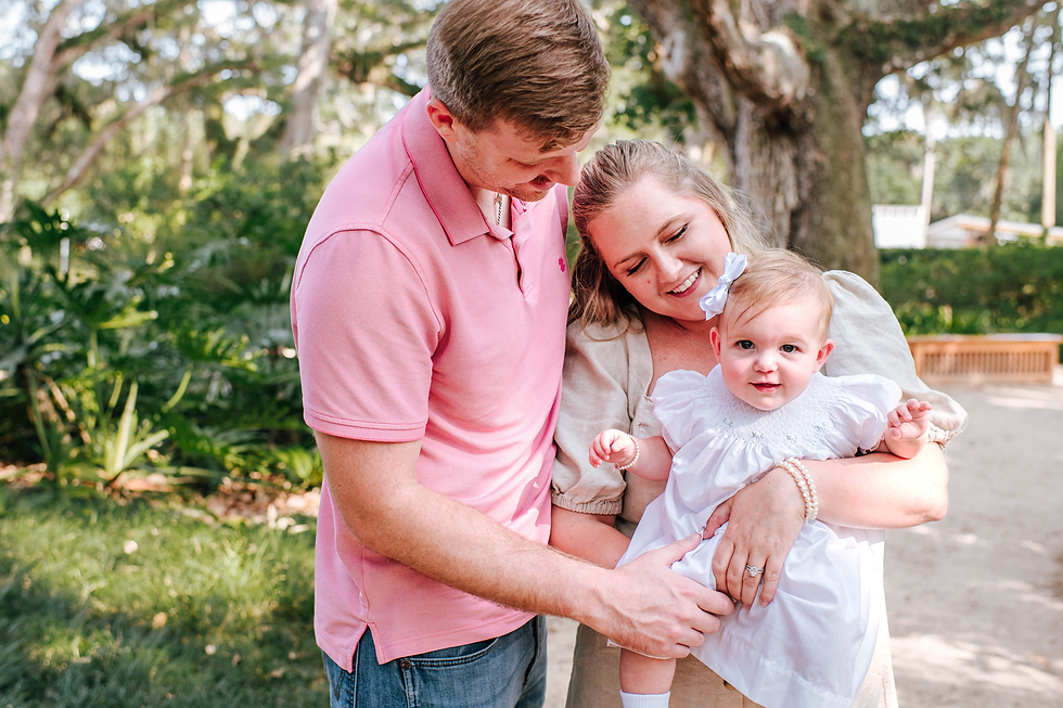 A couple and baby in white dress share a tender moment outside. The man in pink looks down lovingly. Trees and greenery in background making the perfect setting for a family photoshoot.