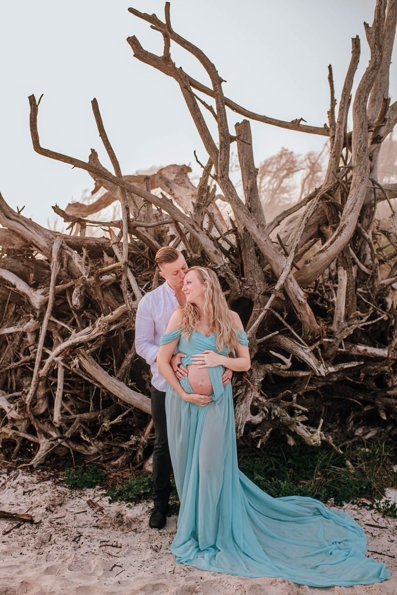 Pregnant couple embraces on a sandy beach with driftwood in the background. Woman in a flowing blue dress, creating a serene atmosphere.