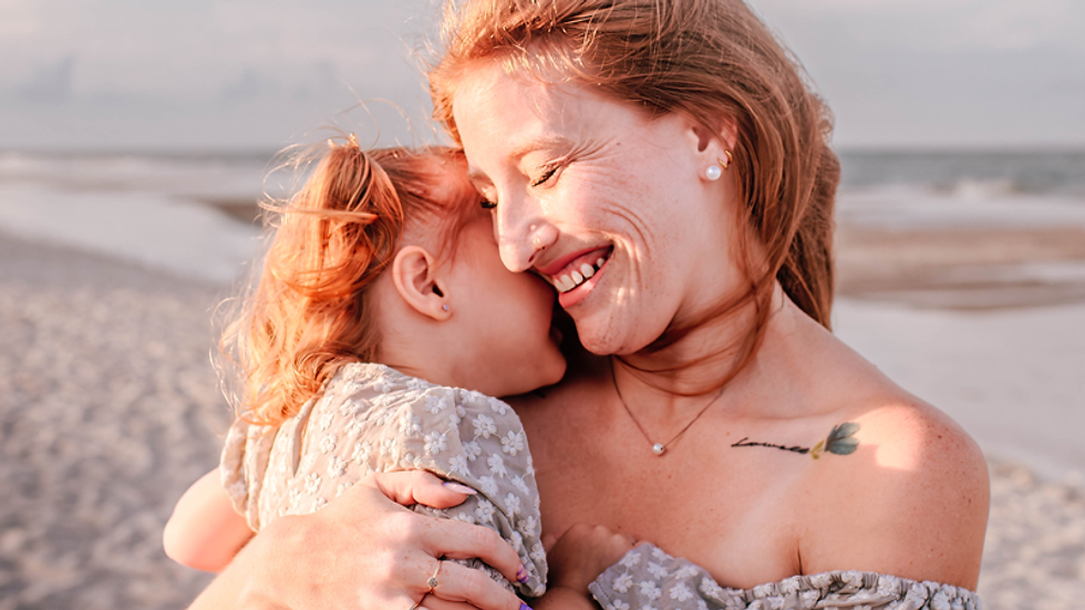 A woman holds a child on a sandy beach at sunset, both wearing floral dresses. They share a joyful, intimate moment under a blue sky.
