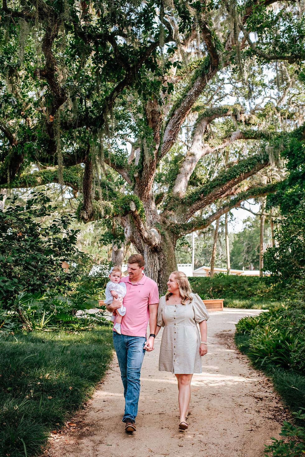 A couple walks on a garden path holding a baby. They are smiling under a large tree with lush green foliage for family photos. Bright and cheerful setting.