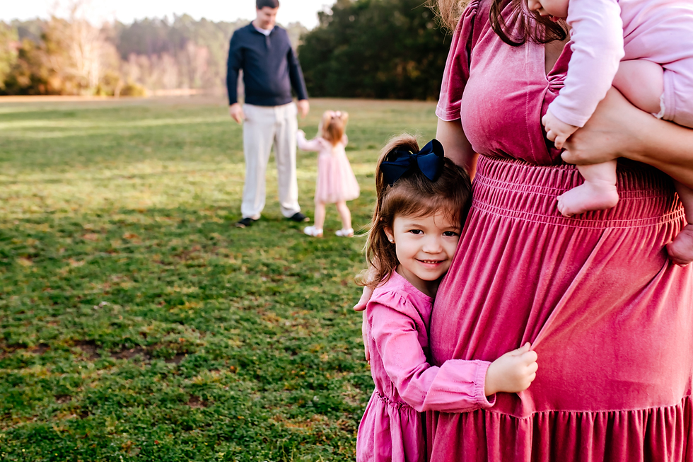 little girl smiles at the local family photographer while hugging her mother who is holding her baby sister.