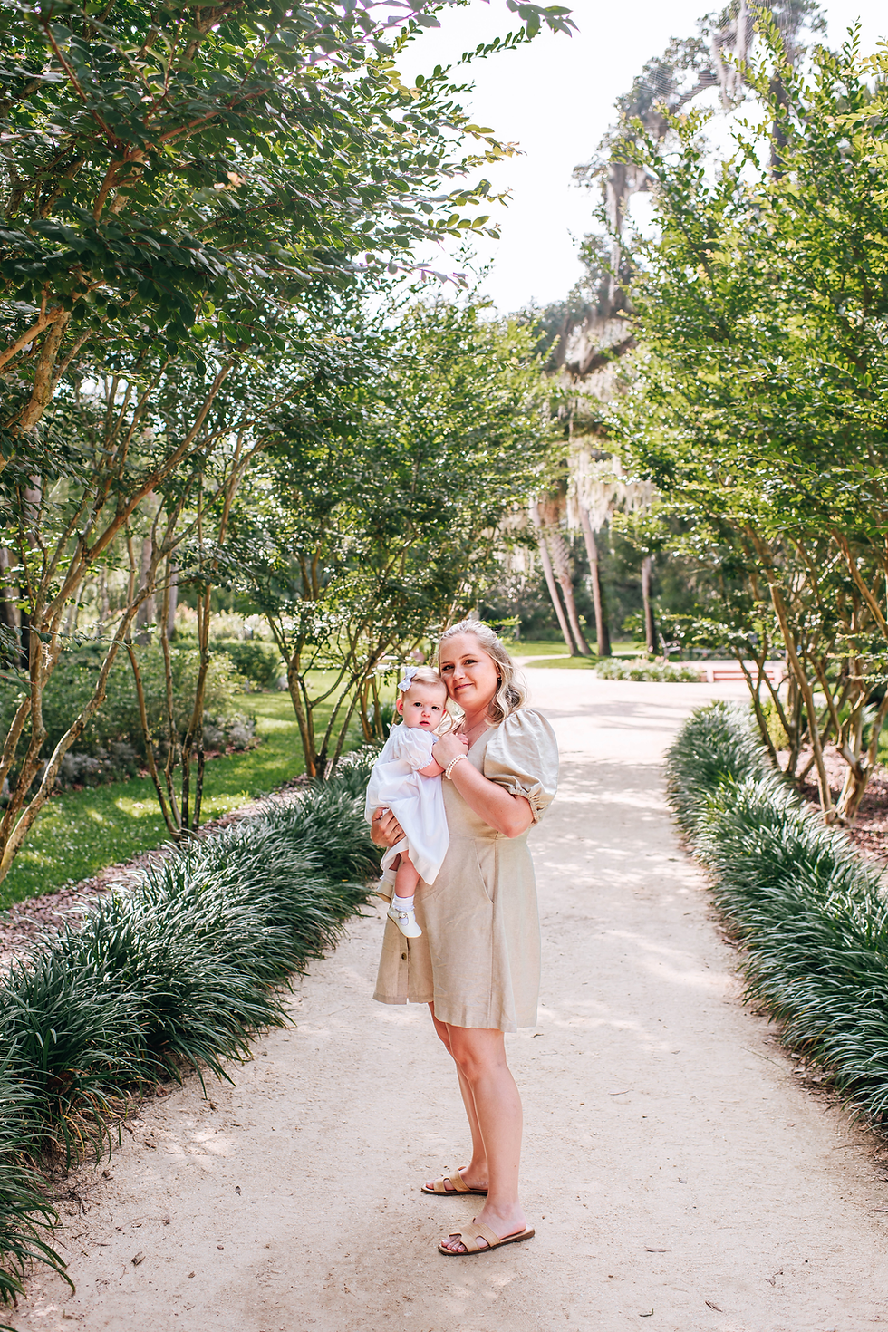 A woman in a light dress holds a baby in a park. They stand on a sunlit path lined with lush green trees and plants, conveying a serene mood during their recent milestone photoshoot.