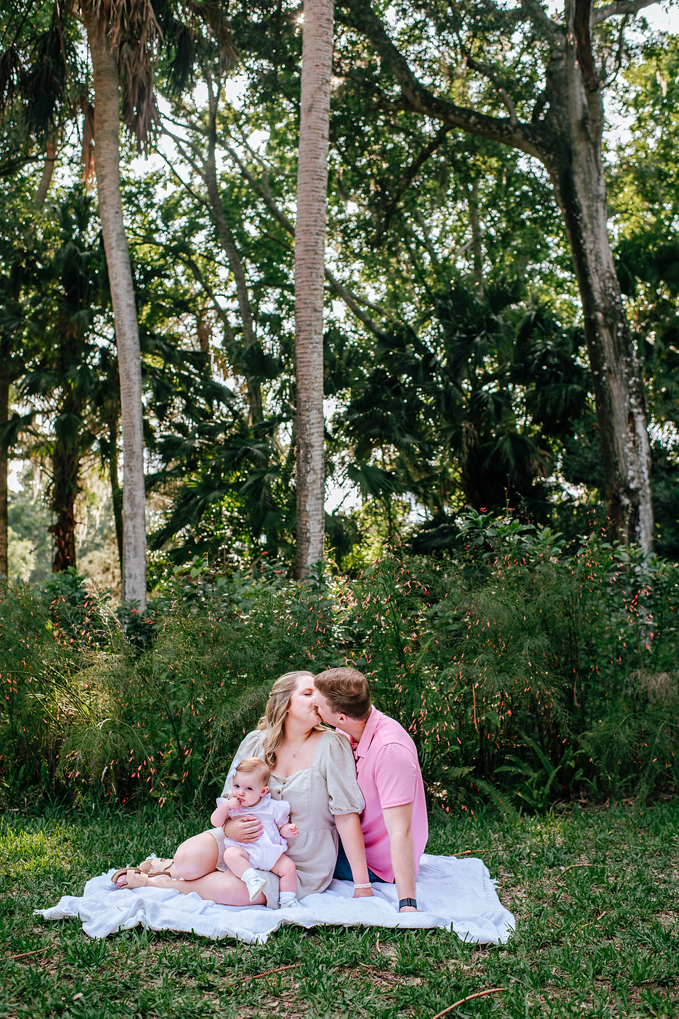 Couple kissing, seated on a blanket with a baby in a lush, green forest. Natural light filters through the trees, creating a serene mood for family photos.