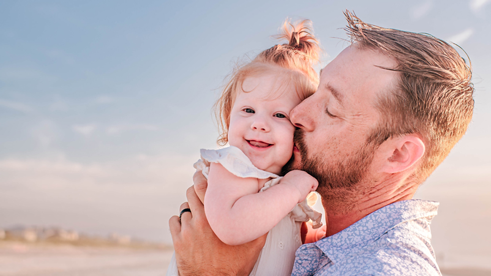 Father kisses smiling baby on cheek at the beach, under a clear blue sky. The man wears a patterned blue shirt, conveying warmth.