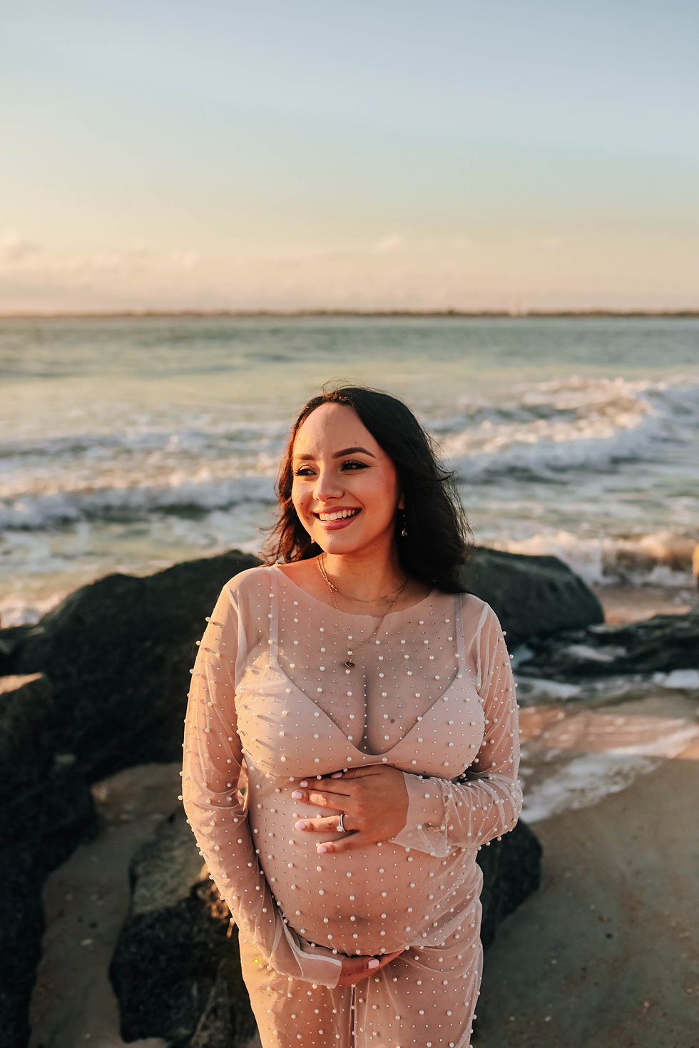 expectant mother laughing as she cradles her tummy during sunrise beach maternity photoshoot
