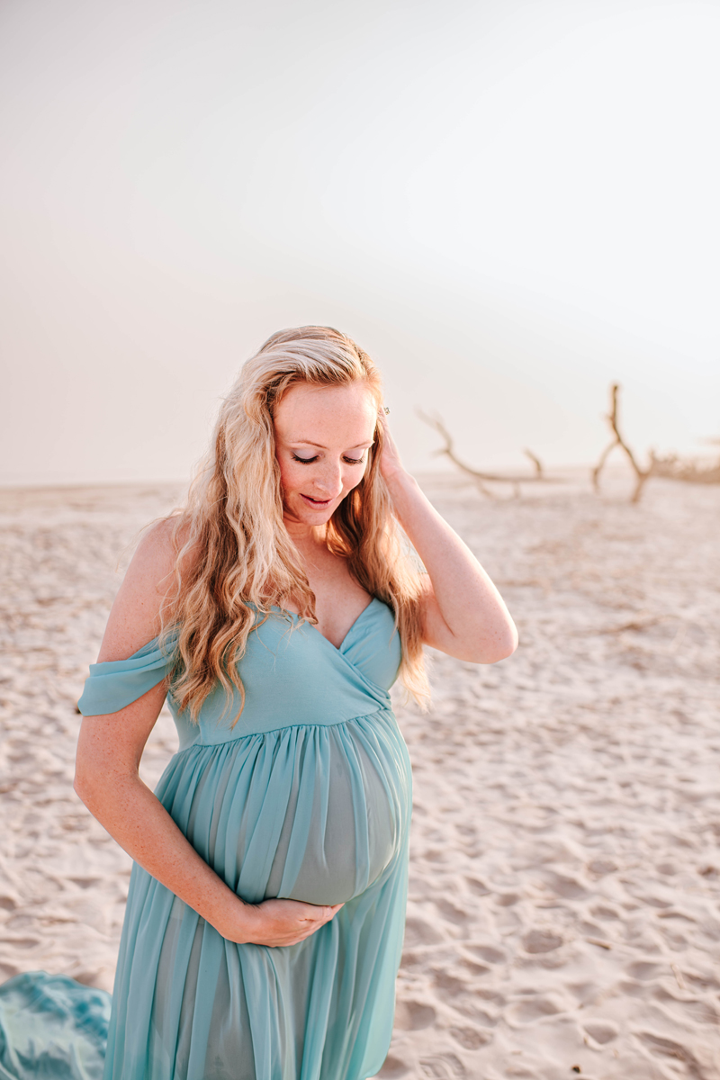 Pregnant woman in a flowing teal dress stands on a sandy beach, gently touching her belly. The sunny setting is peaceful and serene.