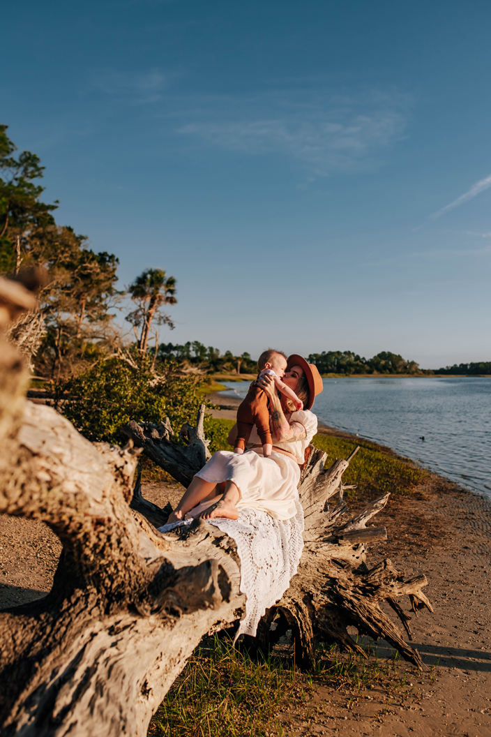 mother holding up baby boy and kissing him while sitting on driftwood tree