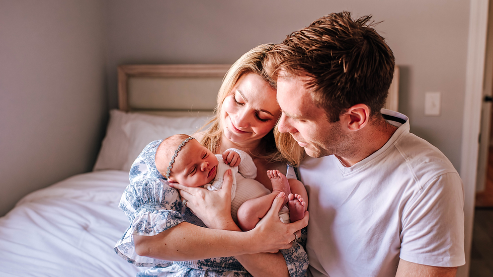 A couple lovingly cradles a newborn in a bedroom with white bedding. The baby wears a headband, and the mood is warm and tender for their in-home newborn photos in Jacksonville, FL.