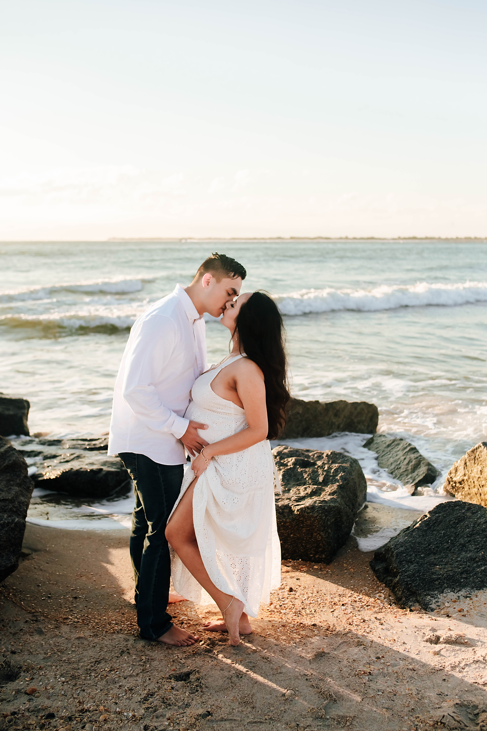 couple kissing on rocky beach at sunrise during maternity photoshoot