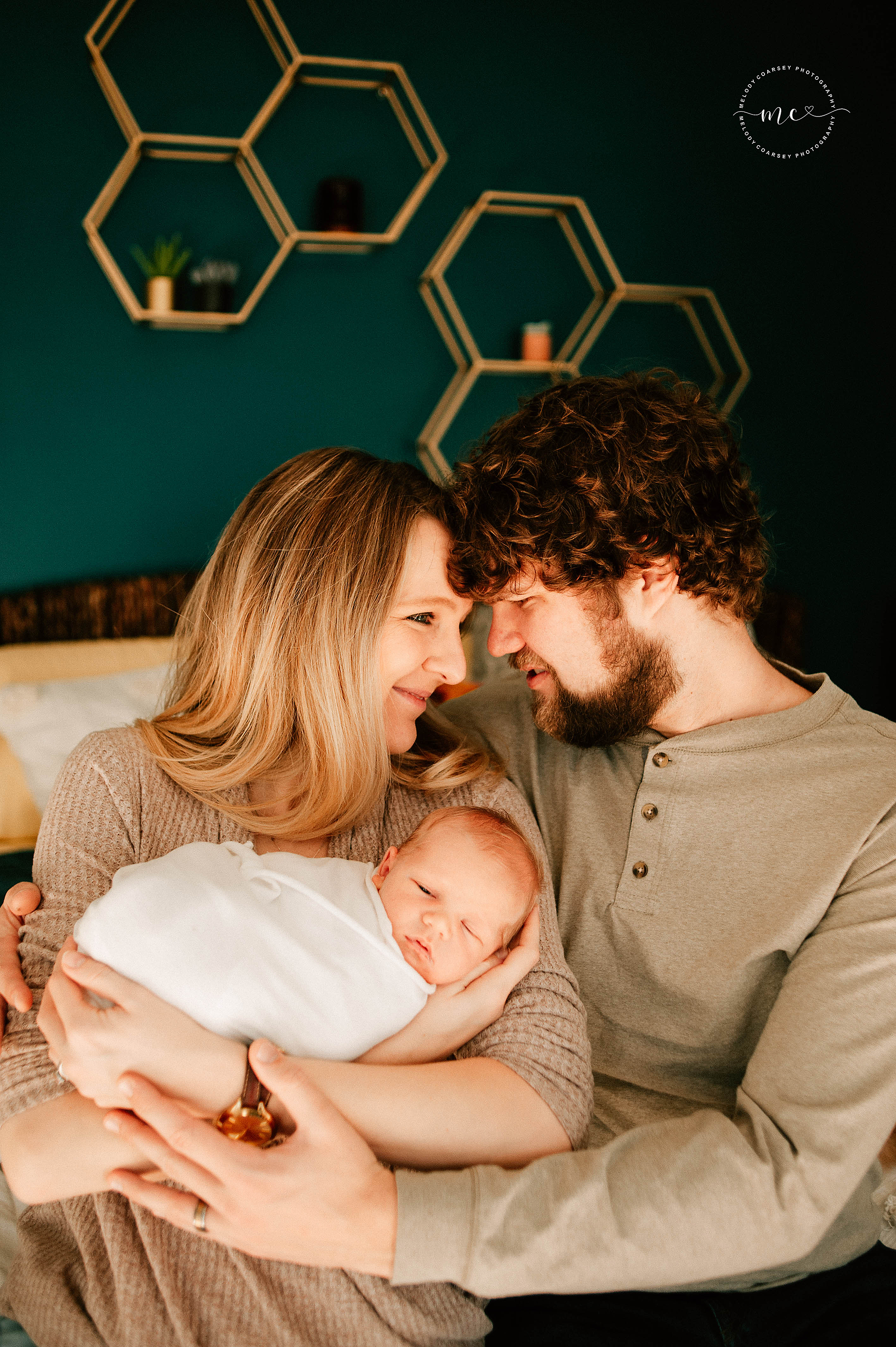 parents touch heads while mother holds newborn swaddled in white in front of green wall