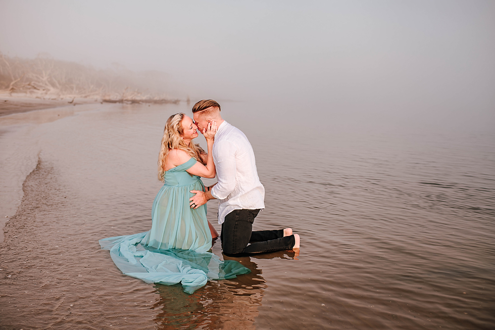 Couple kneeling in shallow water, touching foreheads. Woman in teal dress, man in white shirt. Foggy shoreline, romantic atmosphere.