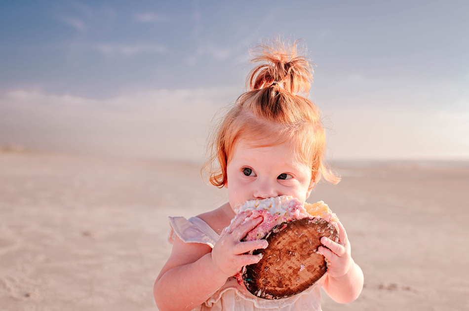 Red-haired toddler eating a colorful smash cake on the beach during a Jacksonville family photography session at sunrise.