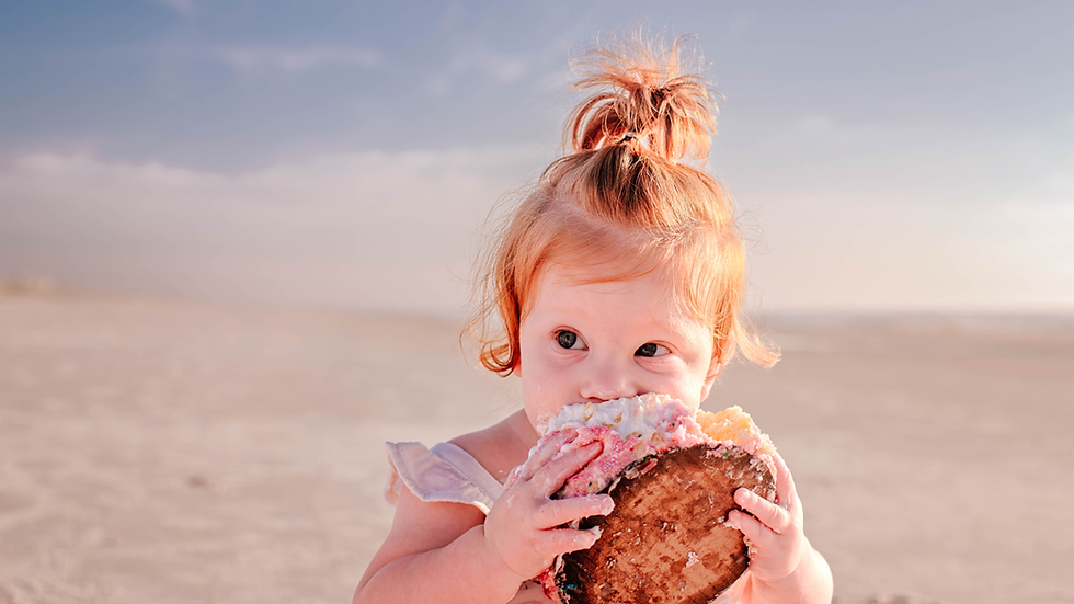 Toddler with red hair joyfully eats her first birthday smash cake on a sandy beach. Bright sky and soft colors create a cheerful, playful mood.