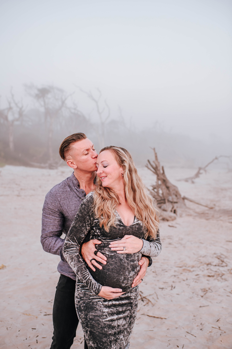 A couple stands on a foggy beach; the man kisses the woman’s head while cradling her baby bump during maternity pictures. She smiles, wearing a velvet dress.