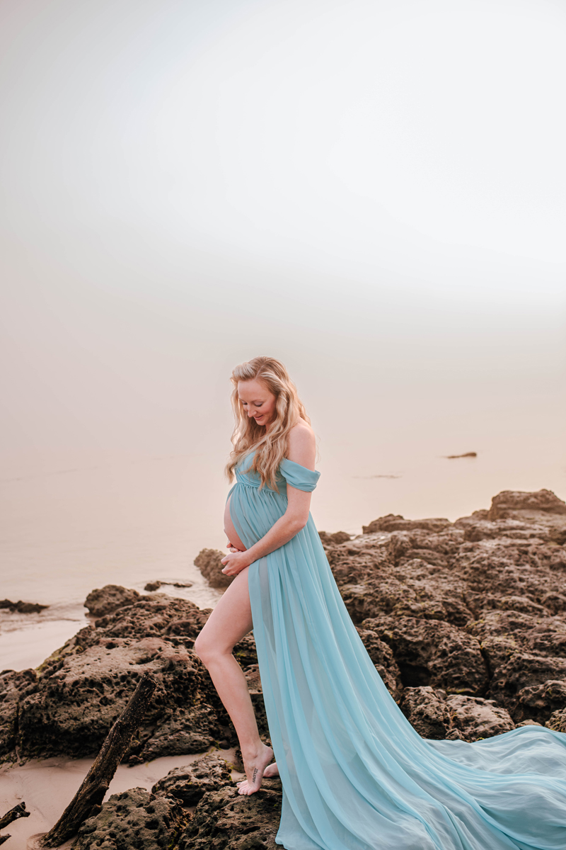 Pregnant woman in a flowing blue dress stands on rocky beach at sunrise, touching her belly and smiling, serene and peaceful mood.