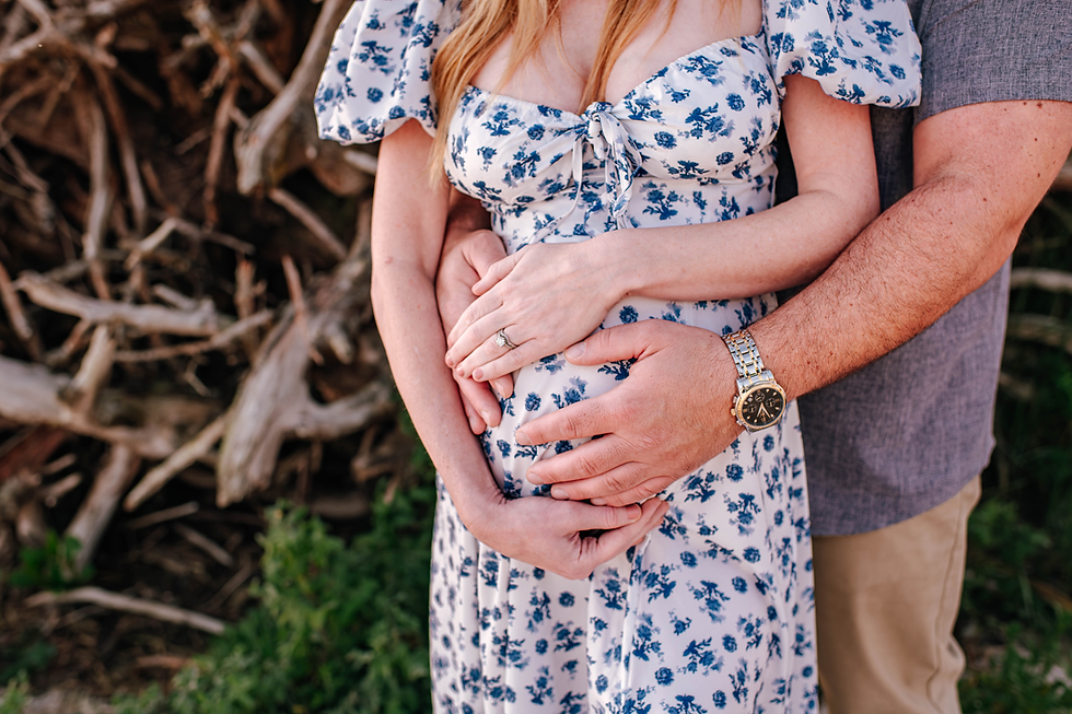 Hands gently cradle a pregnant belly. The woman wears a white dress with blue flowers. The background features rustic wood and greenery.