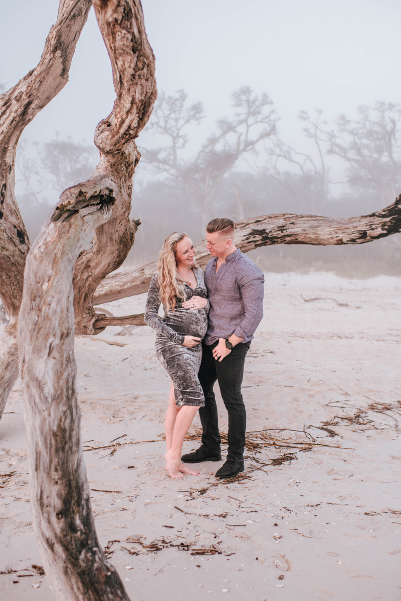 Couple on a foggy beach, standing under twisted driftwood. The woman, in a gray dress, holds her bump, smiling at the man in a blue shirt.