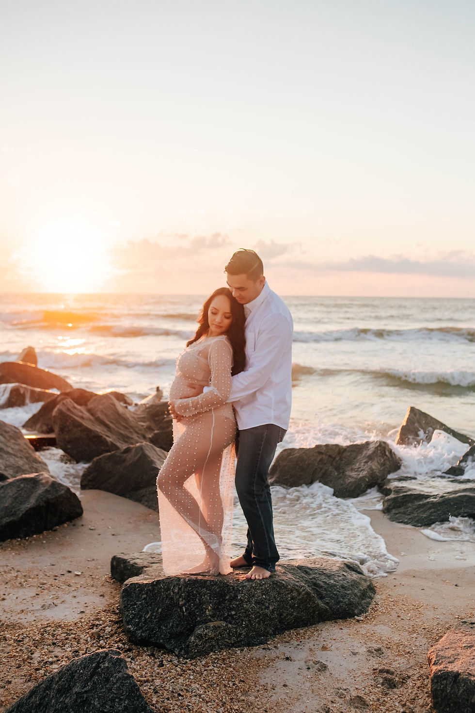 couple standing on rocky beach at sunrise for maternity photos