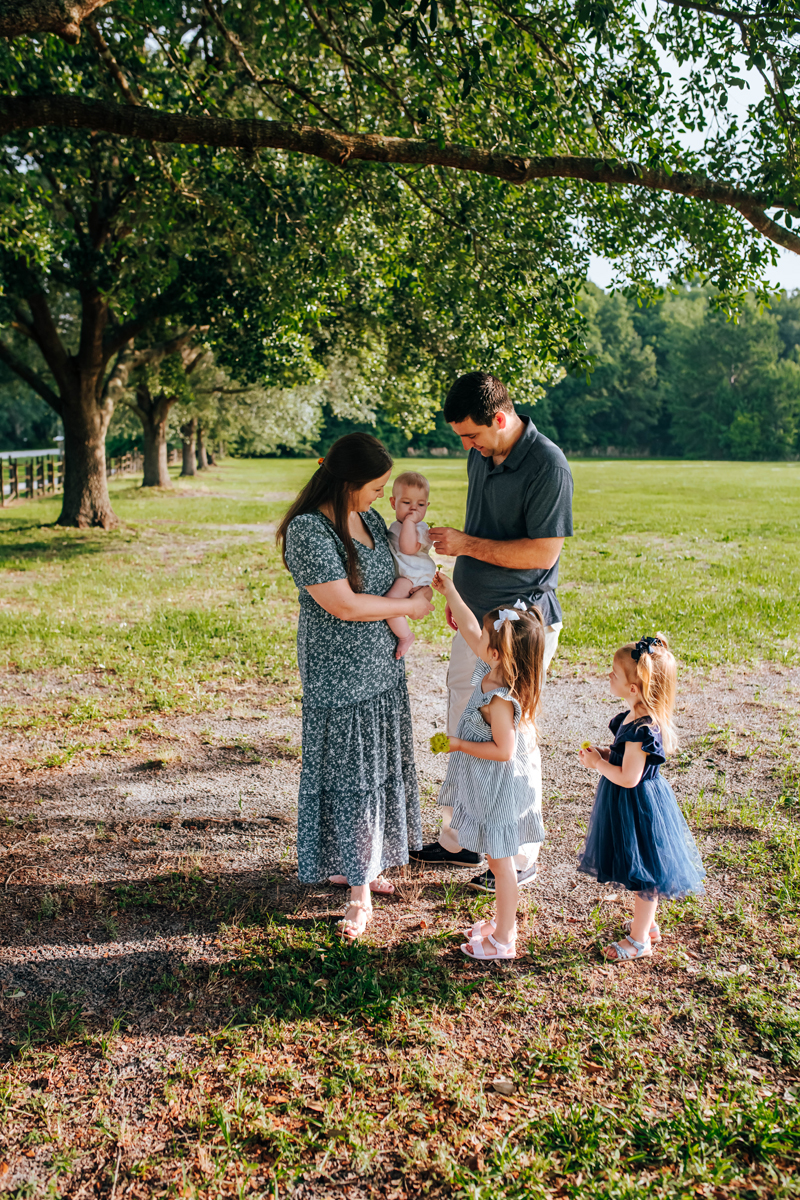 parents hold baby girl as older daughters bring flowers they picked during a park family photoshoot