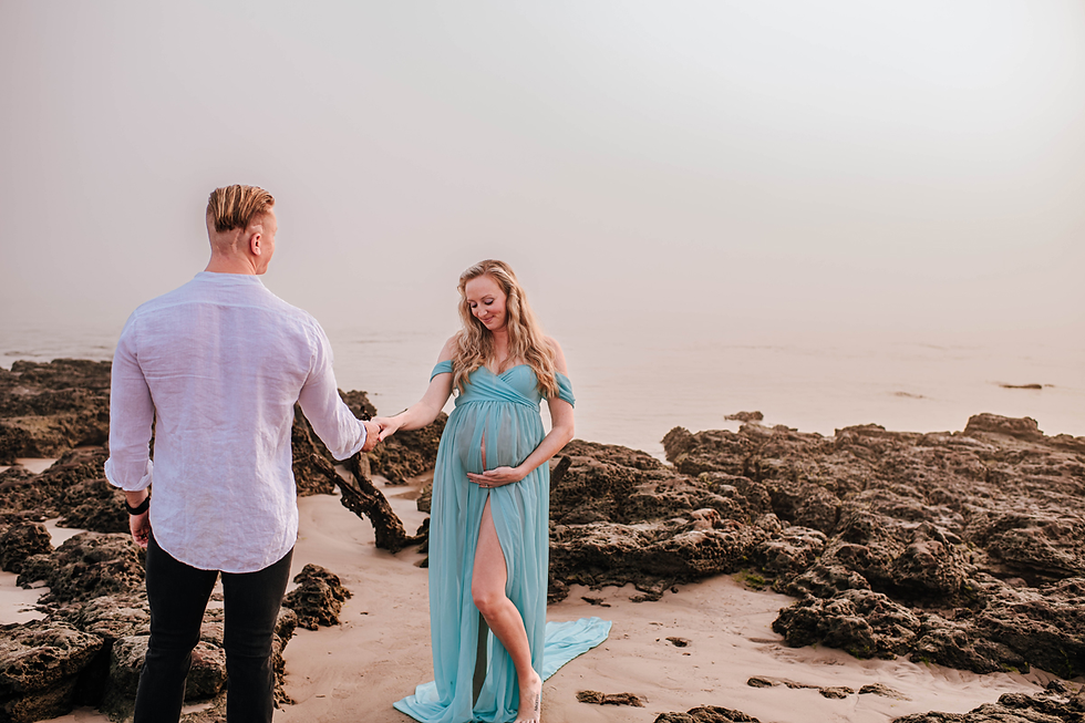 Man and pregnant woman holding hands on a rocky beach. She wears a flowing blue dress, caressing her belly, with a serene expression.