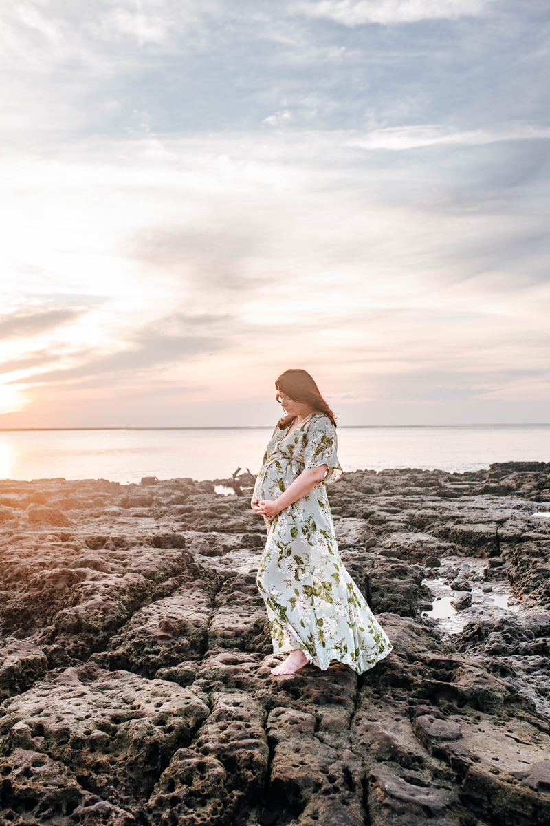 pregnant woman in a green floral dress stands on the rocks with the ocean sunrise behind her posing for maternity photoshoot