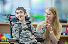 Young male boy with brown hair and light skin sitting in a wheelchair next to a blonde hair, light skin female. Both are smiling and female is holding males hand. Male has black chest harness across chest and a red headswitch positioned to his right headrest.