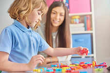 Young boy with blonde hair, light skin and a blue shirt picking up letter magnets with a brunette, light skin woman at a table.
