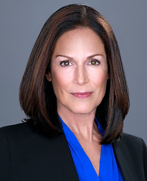 Corporate headshot of white woman with brown hair wearing blue shirt and black blazer in front of grey background