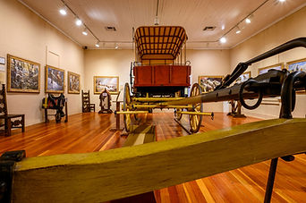 A low angle image of a replica of a 19th century covered wagon. It is in the interior of a building, and mounted on the surrounding walls are numerous oil paintings.
