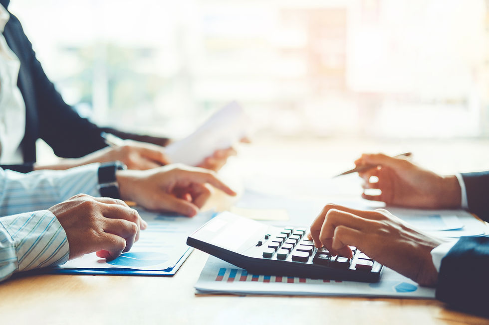 Hands working at a table with papers, graphs, and a calculator. People in business attire, bright office setting, and focused mood.