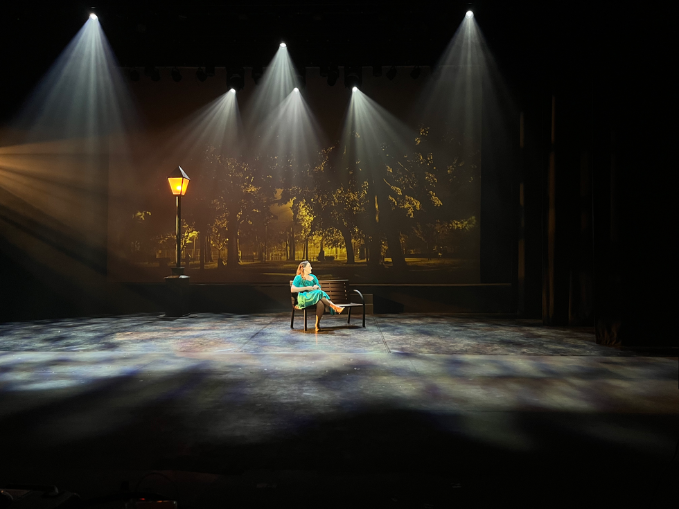 An actor sat on a bench with scenic lighting and video surrounding her.