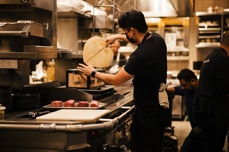 Chef tending to the grill with a hand fan beside thick cuts of steak, showcasing QUA’s liv