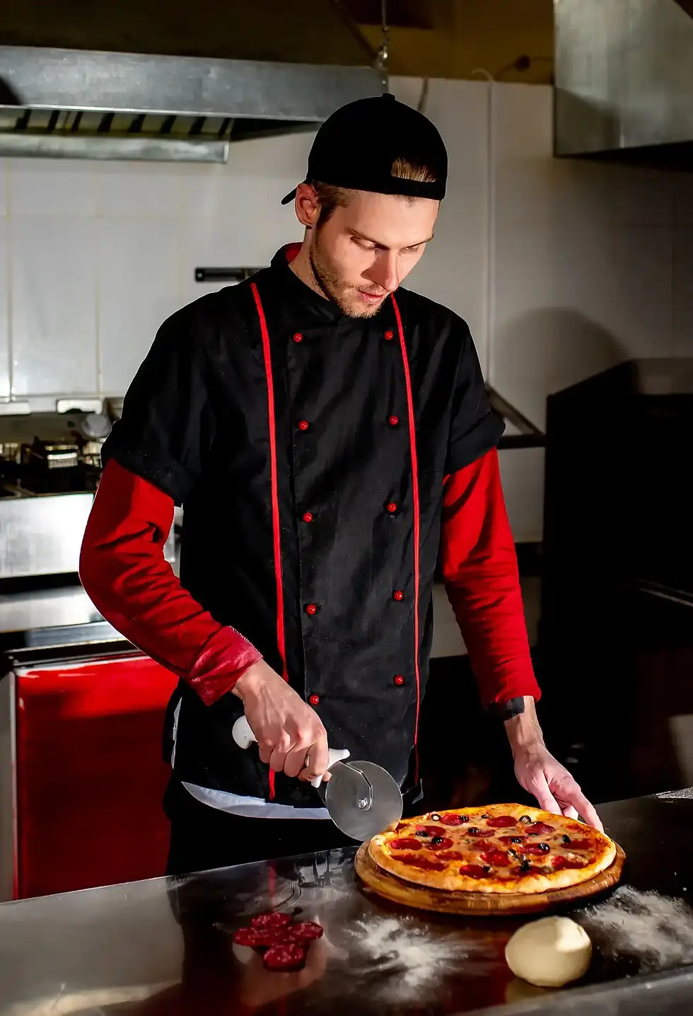 Man in uniform cutting a pepperoni pizza in a commercial kitchen.
