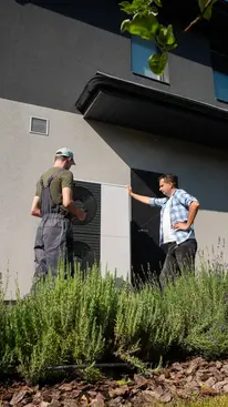 Two men standing near a building discussing something outdoors with plants.