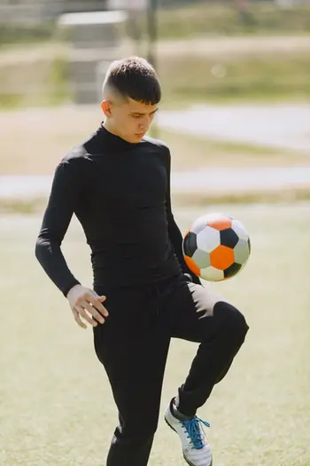 Young boy wearing black plays soccer; holding ball on foot; outdoors field.