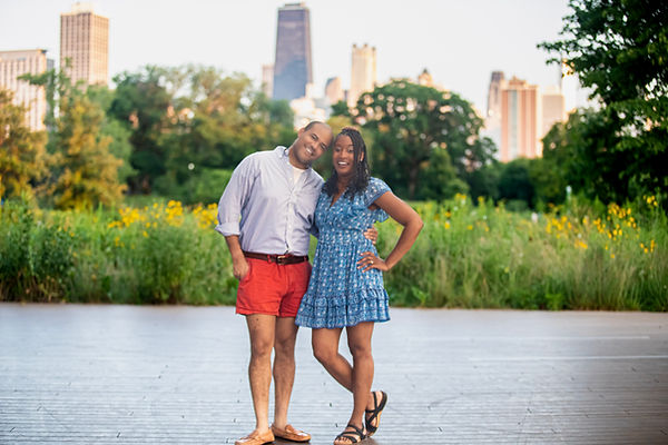 Chicago engagement photo of couple standing barefoot in a grassy field with skyline in background, woman in blue dress and man in red shorts smiling together at sunset
