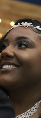 Bride in a white wedding gown holds onto groom’s arm as they stand together in an elegant indoor portrait with soft window light and dark interior background.